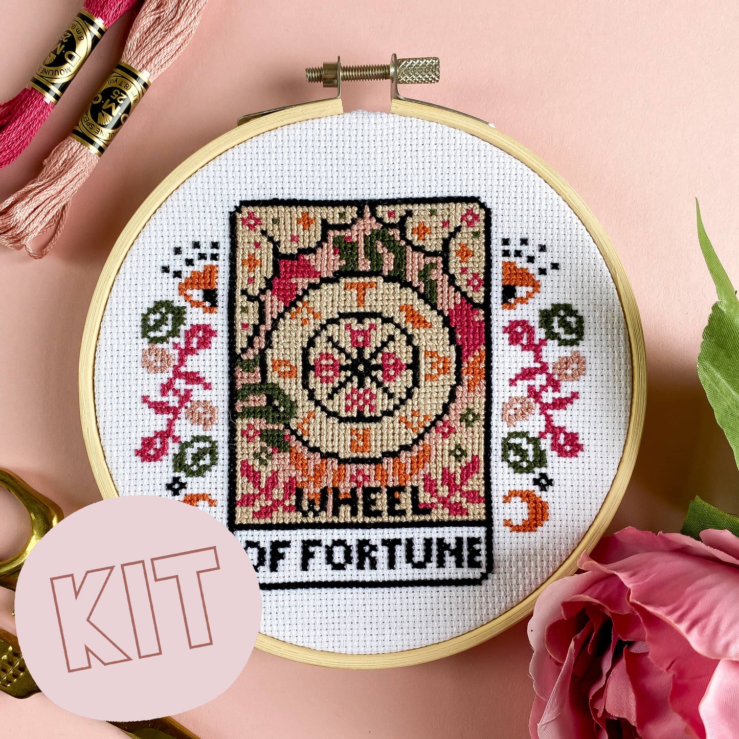 Embroidery hoop with cross-stitched design of 'Wheel of Fortune' on a pink background.