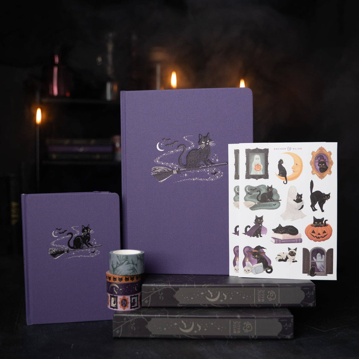 Purple stationery set featuring a large and small hardcover notebook, a sheet of Halloween-themed stickers, and a roll of washi tape, styled on dark books with warm candlelight in the background.