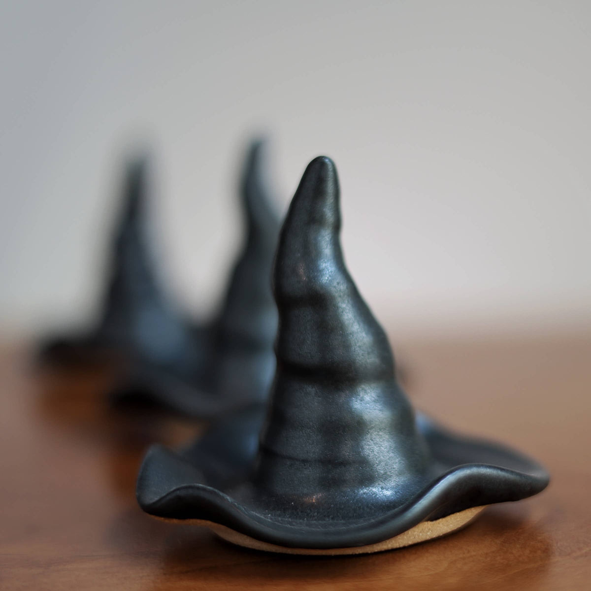 Three small black witch hat-shaped objects arranged in a row on a wooden surface, with the front one in sharp focus and the others blurred in the background against a soft neutral backdrop.