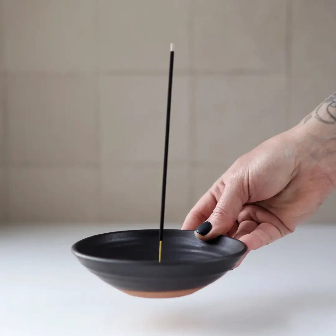 Hand holding a black incense holder with a single incense stick against a neutral background