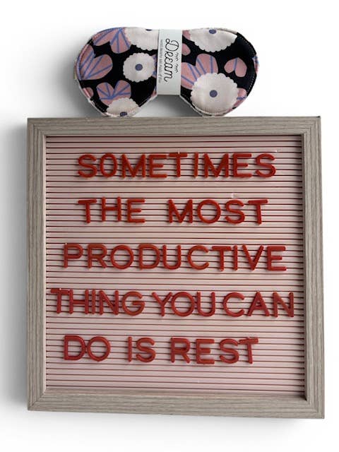A framed letter board sign with red letters reading 'SOMETIMES THE MOST PRODUCTIVE THING YOU CAN DO IS REST' on a light pink background, with a matching floral sleep mask above it.