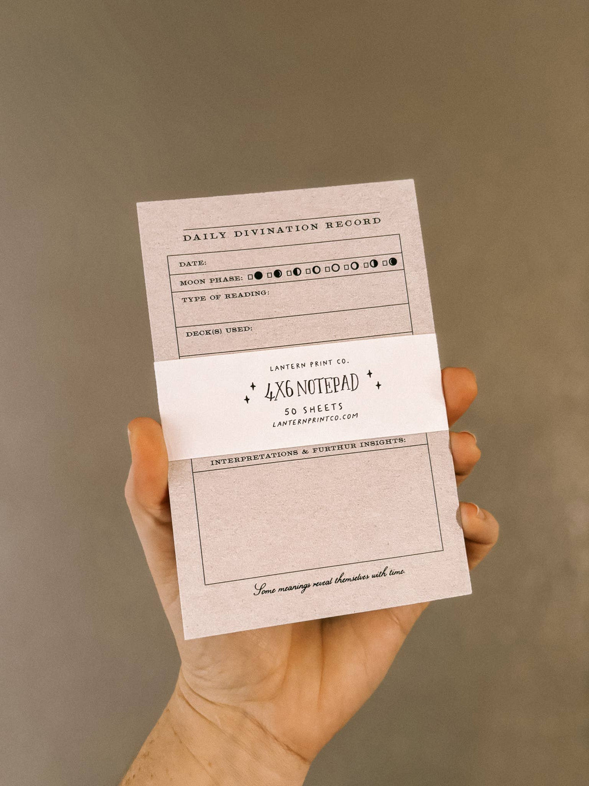 A hand holding a 4x6 notepad with a white paper band across the middle. The notepad has a textured light beige paper background with black printed text and lines, including a 'Daily Divination Record' section.