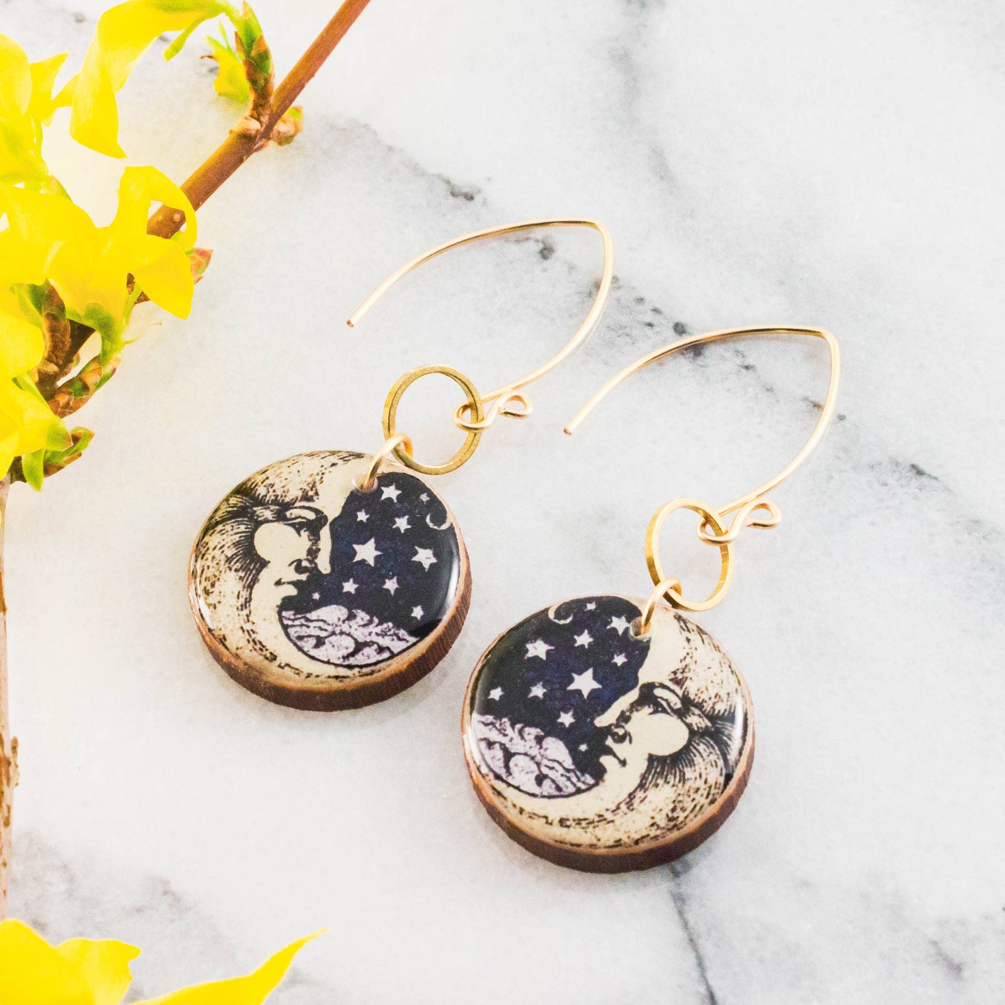 Pair of earrings with moon and star design on a marble surface with yellow flowers.
