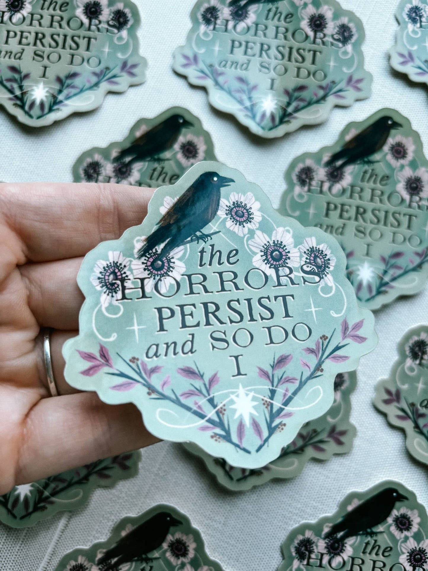 Sticker with 'the Horrors Persist and So Do I' text held by a hand, surrounded by more stickers.