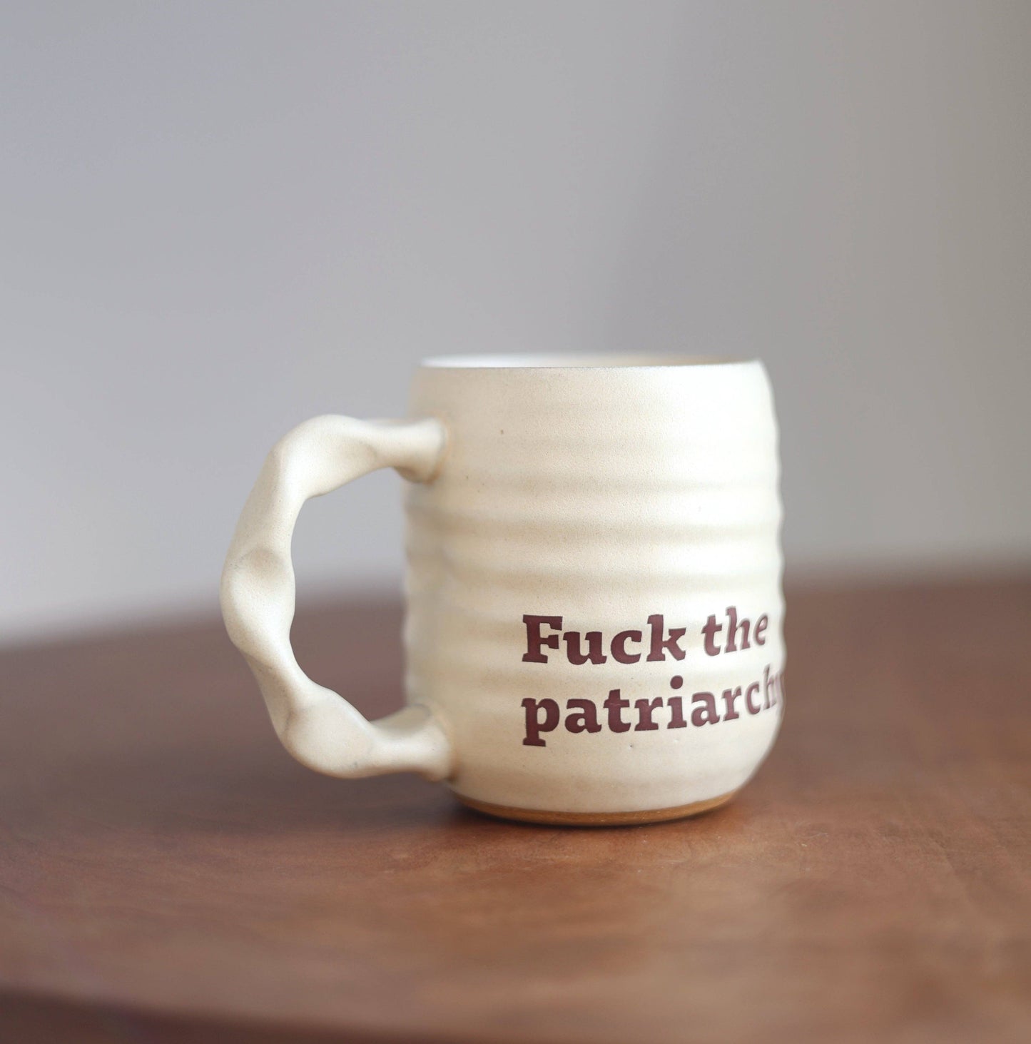 An off-white ceramic mug with a sculptural, wavy handle and the text 'Fuck the patriarchy' printed in dark brown on the side, resting on a wooden surface against a neutral gray background.