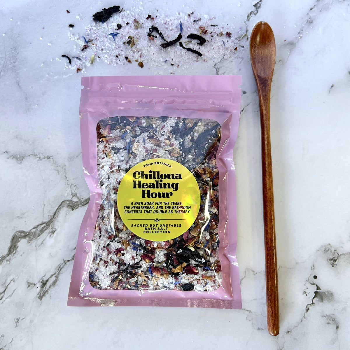 A pink resealable pouch labeled 'Chilpa Healing Hour' by Sacred Earth Vegetables, filled with a multicolored herbal mixture, placed on a white marble surface with a wooden spoon beside it.