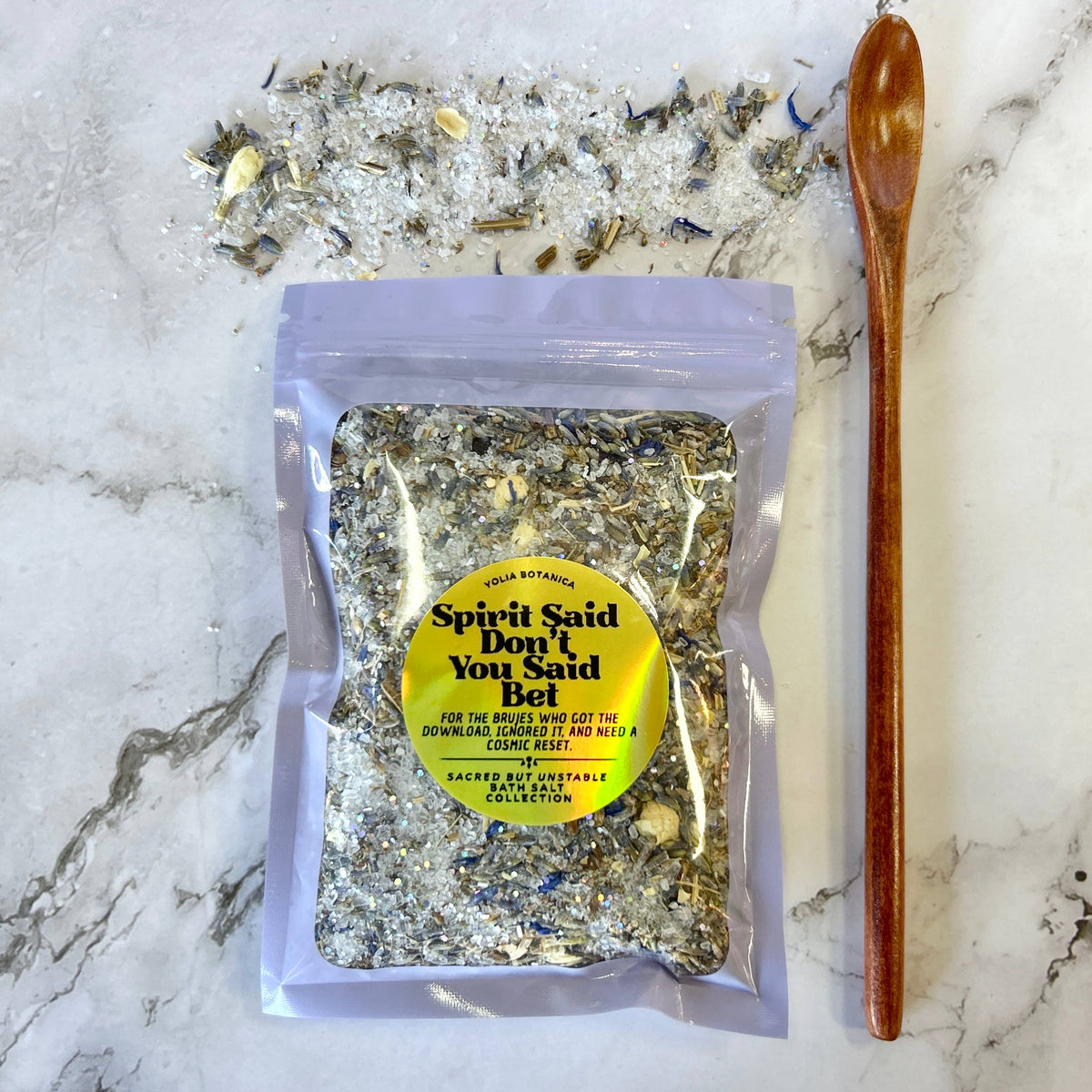 A clear stand-up pouch containing a textured herbal salt blend, placed on a white marble surface with loose salt crystals scattered behind it and a wooden spoon to the right. The pouch has a yellow circular label reading 'Spirit Said Don't You Said Bel' and 'Sage Botanicals'.