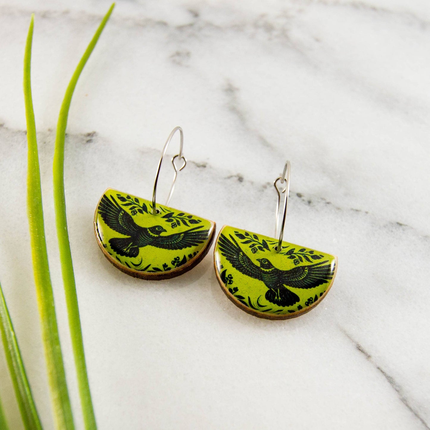 Green earrings with black bird design on a marble surface with green leaves.