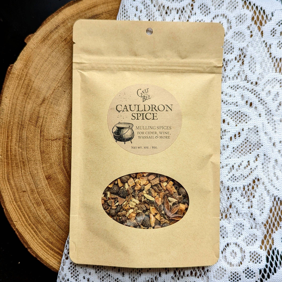A kraft paper stand-up pouch labeled 'Cauldron Spice' with a circular window showing mixed peppercorns and spices, photographed on a wooden slice and white lace cloth background.