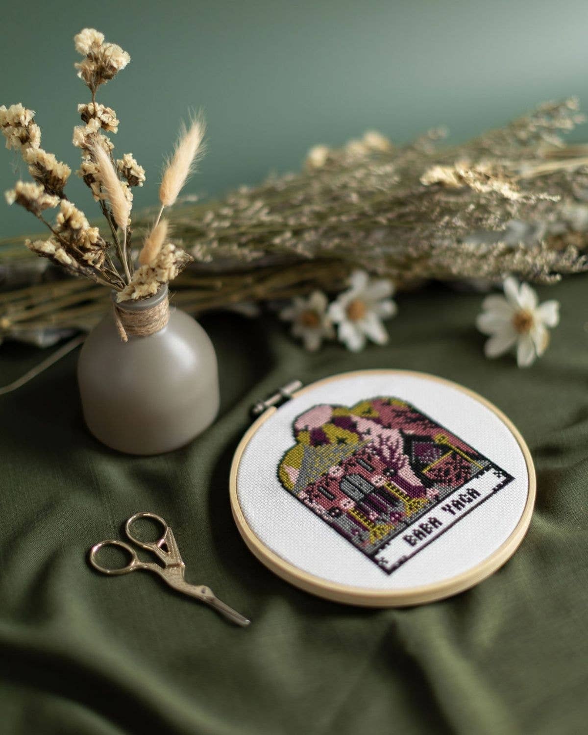 Embroidery hoop with a completed cross-stitch design on a green fabric background with dried flowers and scissors.