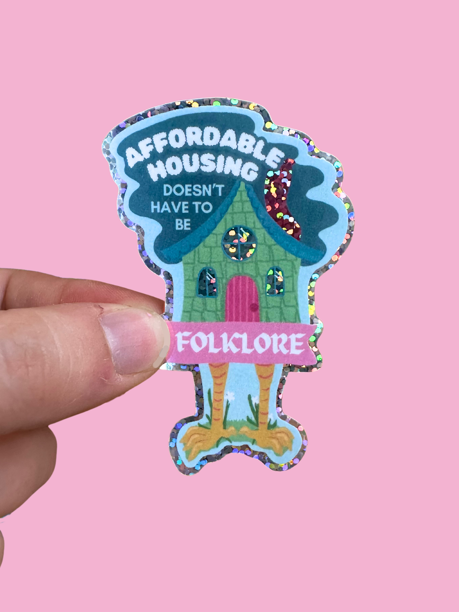 Sticker with Baba Yaga's hut design and text 'Affordable Housing Doesn't Have to Be Folklore' held against a pink background.