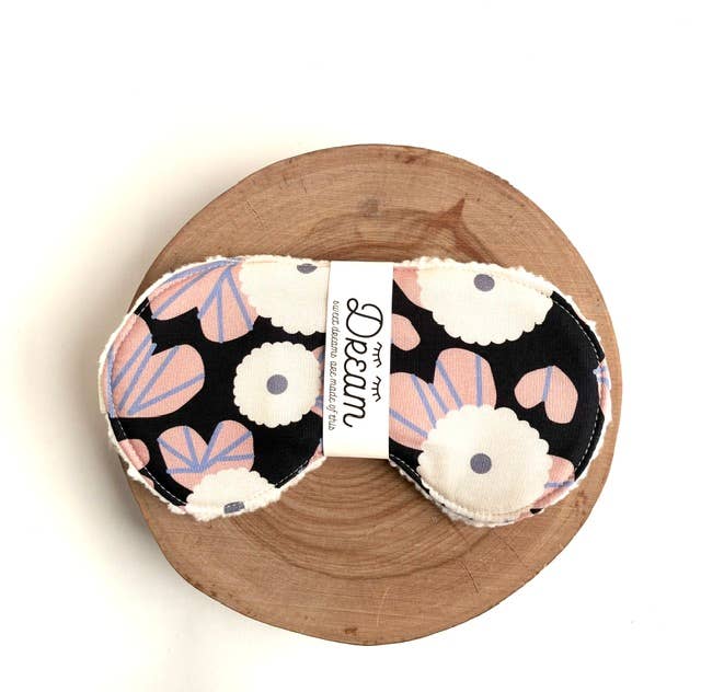 A pair of fabric eye masks with a black background and a floral pattern of white flowers, pink petals, and lavender-blue accents, displayed on a round wooden slice against a white background.