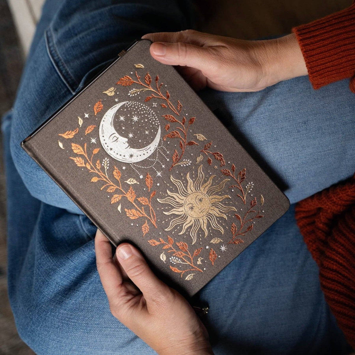 A person holding a dark brown journal with an embossed celestial design of a crescent moon and sun surrounded by autumn leaves, set against blue jeans and a rust-colored sweater.