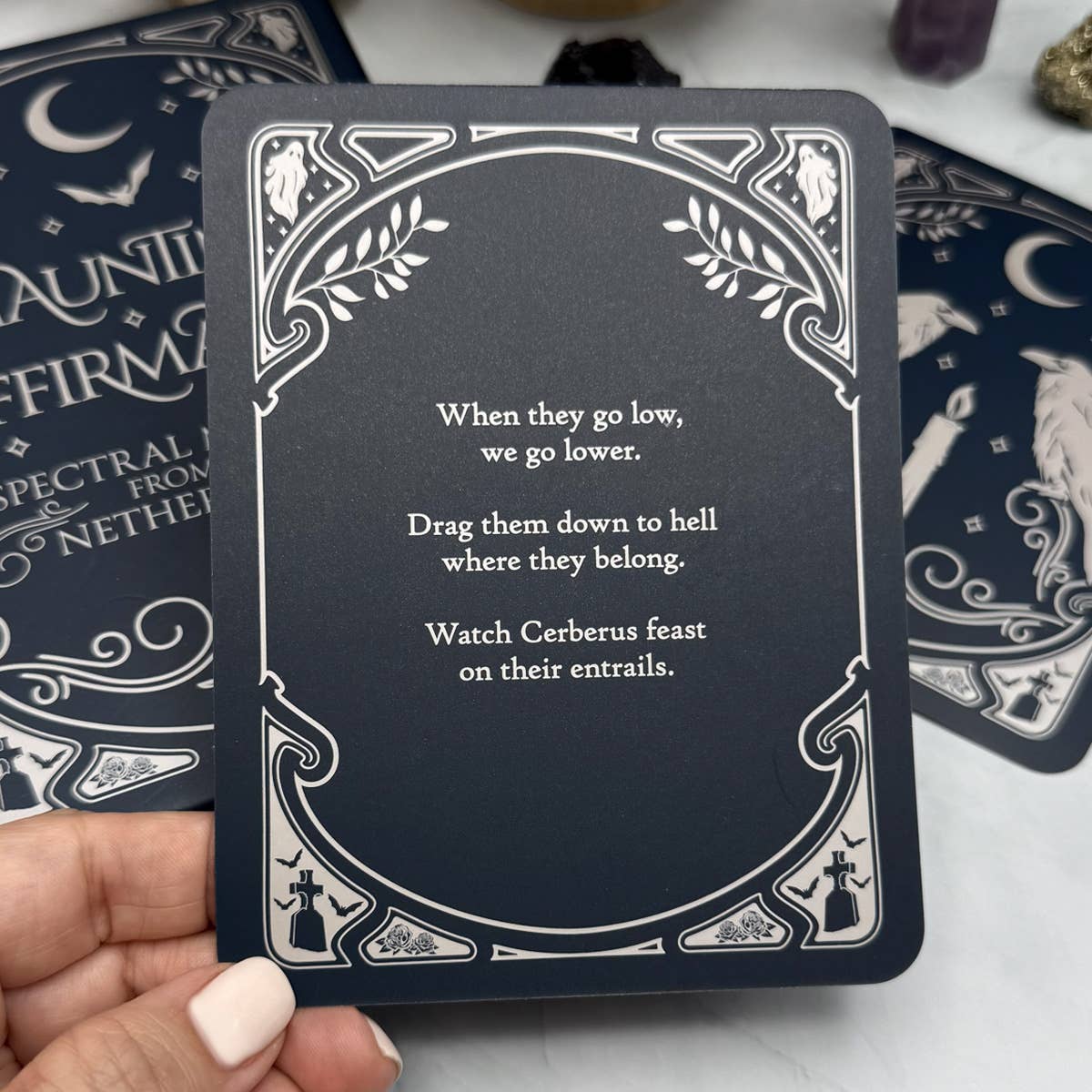 Black card with white text and decorative border held by a hand, with tarot cards in the background.