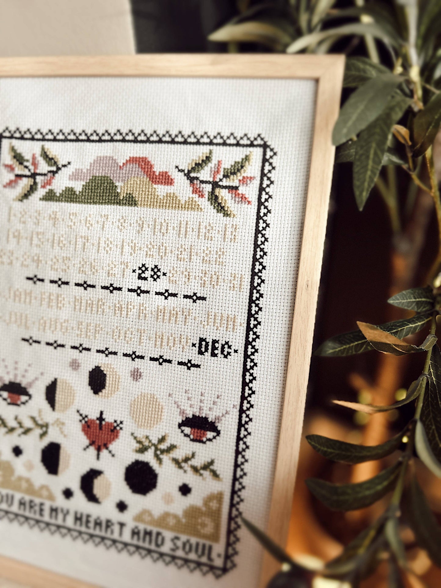 Framed cross-stitched artwork with decorative elements and text, placed on a surface with a plant in the background.
