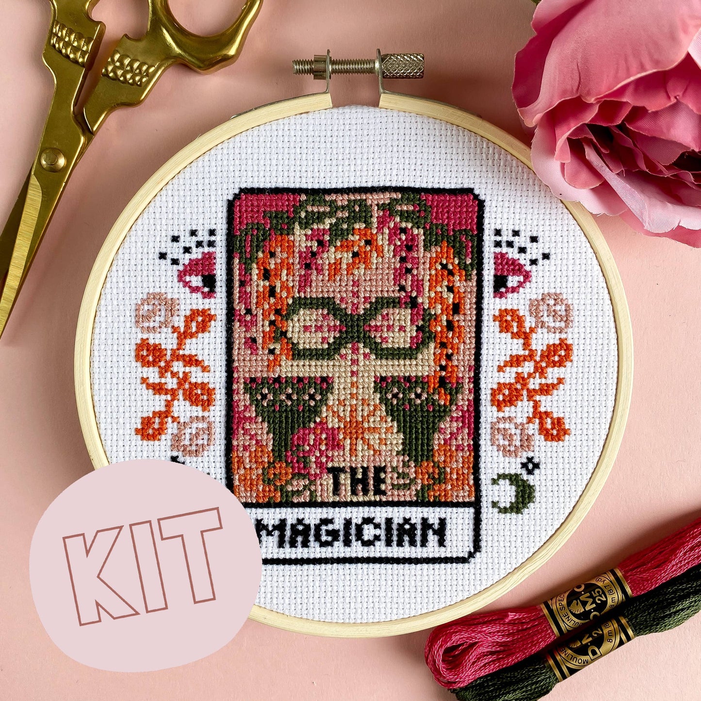 Cross-stitch kit with 'The Magician' tarot card design, surrounded by embroidery tools and pink flowers.