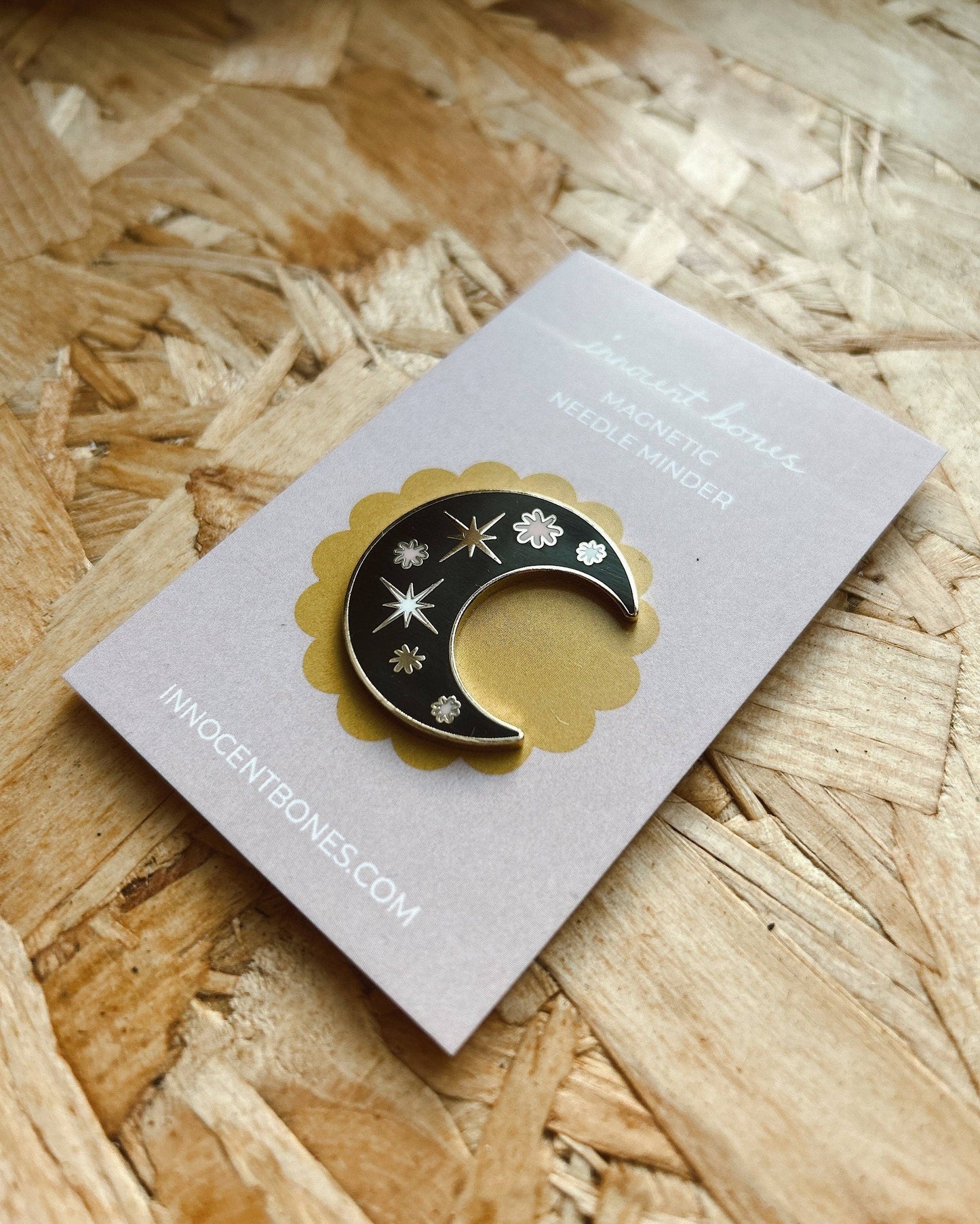 Black and gold crescent moon-shaped pin on a card with 'innocentbones.com' branding, placed on a woven surface.