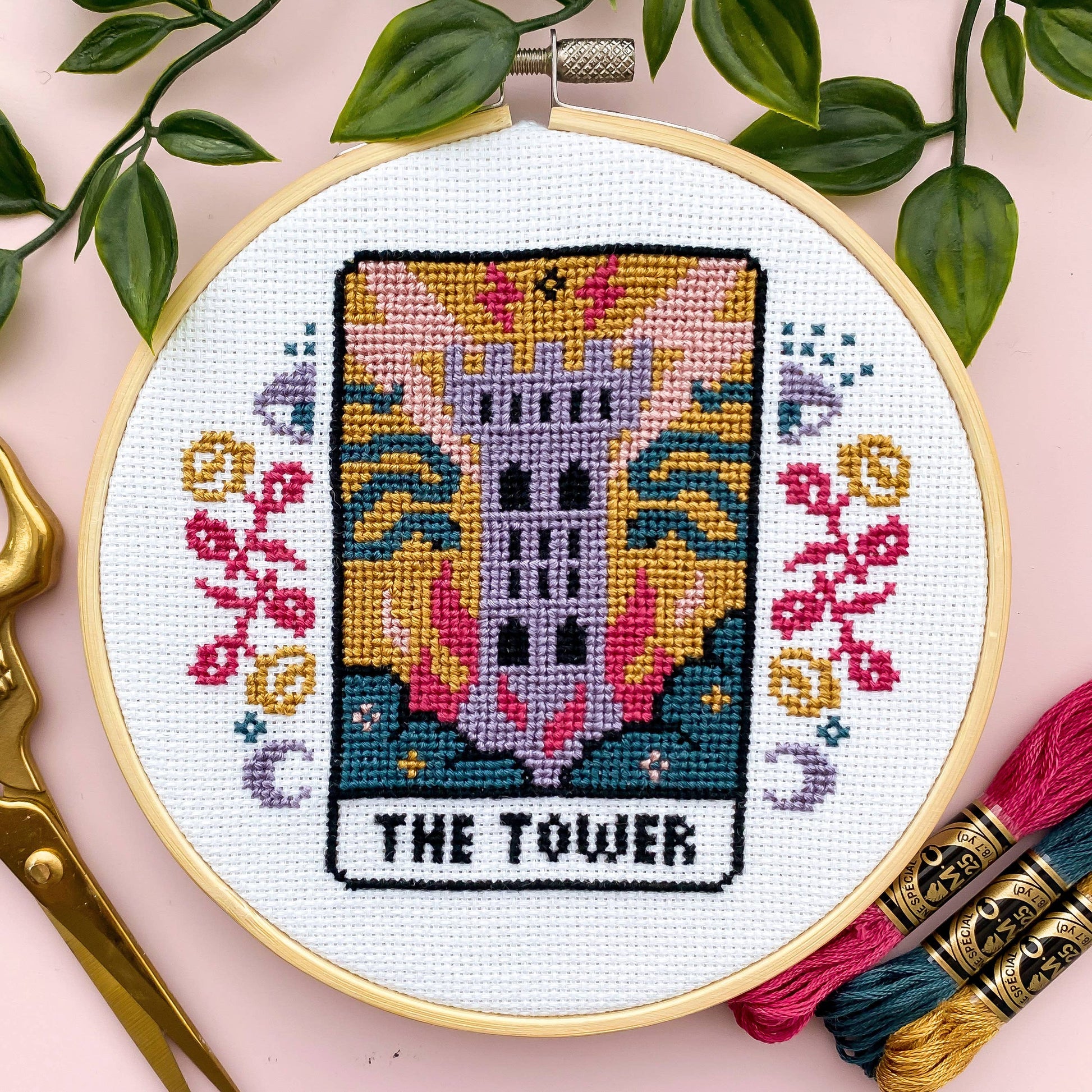 Cross-stitched tarot card design with 'The Tower' on a white fabric background, surrounded by green leaves and embroidery threads.
