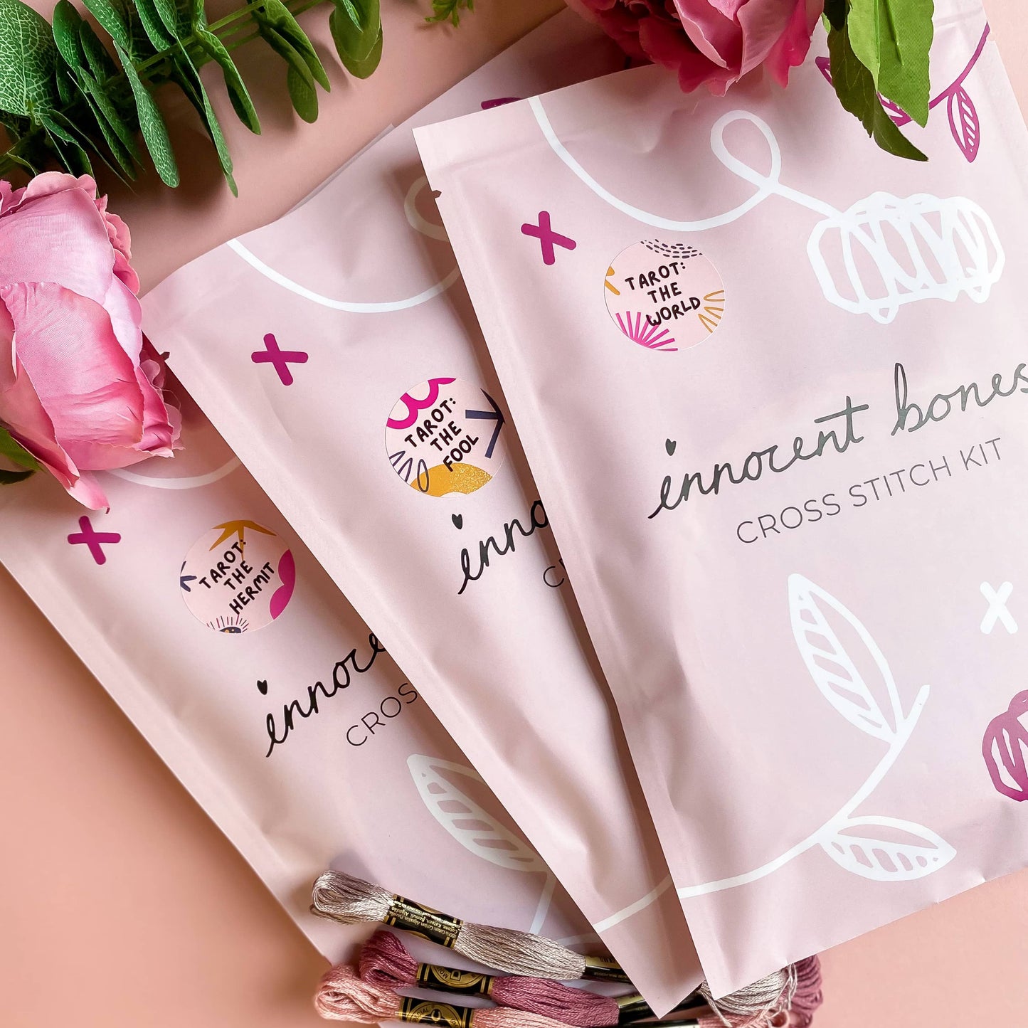 Cross stitch kits with floral designs and text on a pink surface.