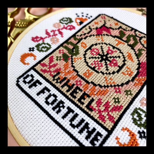Cross-stitched design with 'Wheel of Fortune' text and decorative patterns in an embroidery hoop.