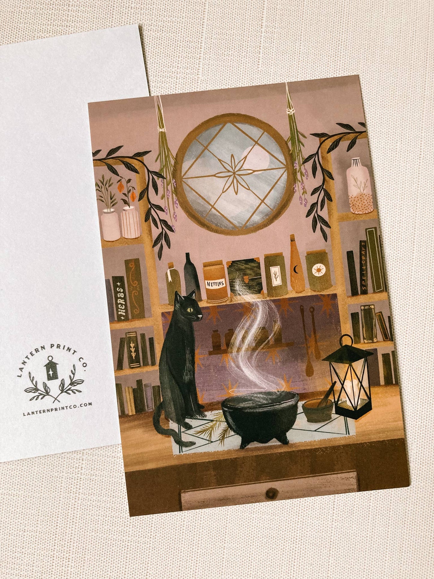 Greeting card with a black cat and witch's cauldron on a bookshelf background, featuring Lantern Press logo.