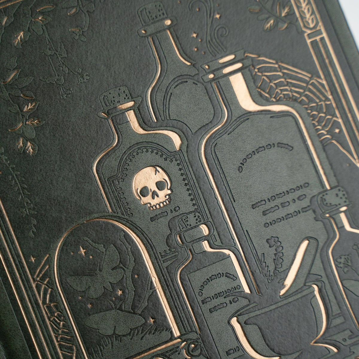 Close-up of a dark textured surface featuring embossed gothic illustrations of potion bottles, a skull, a frog under glass, and spiderwebs in a metallic gold tone.