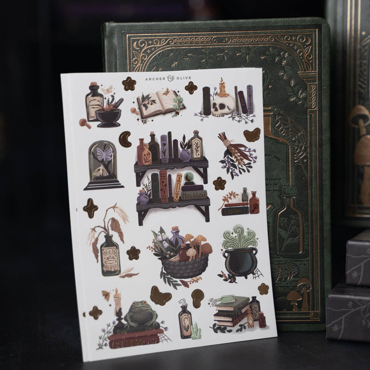 A sheet of decorative stickers featuring a witchy and potion-themed illustration set, displayed in front of a vintage-style green book on a dark background.