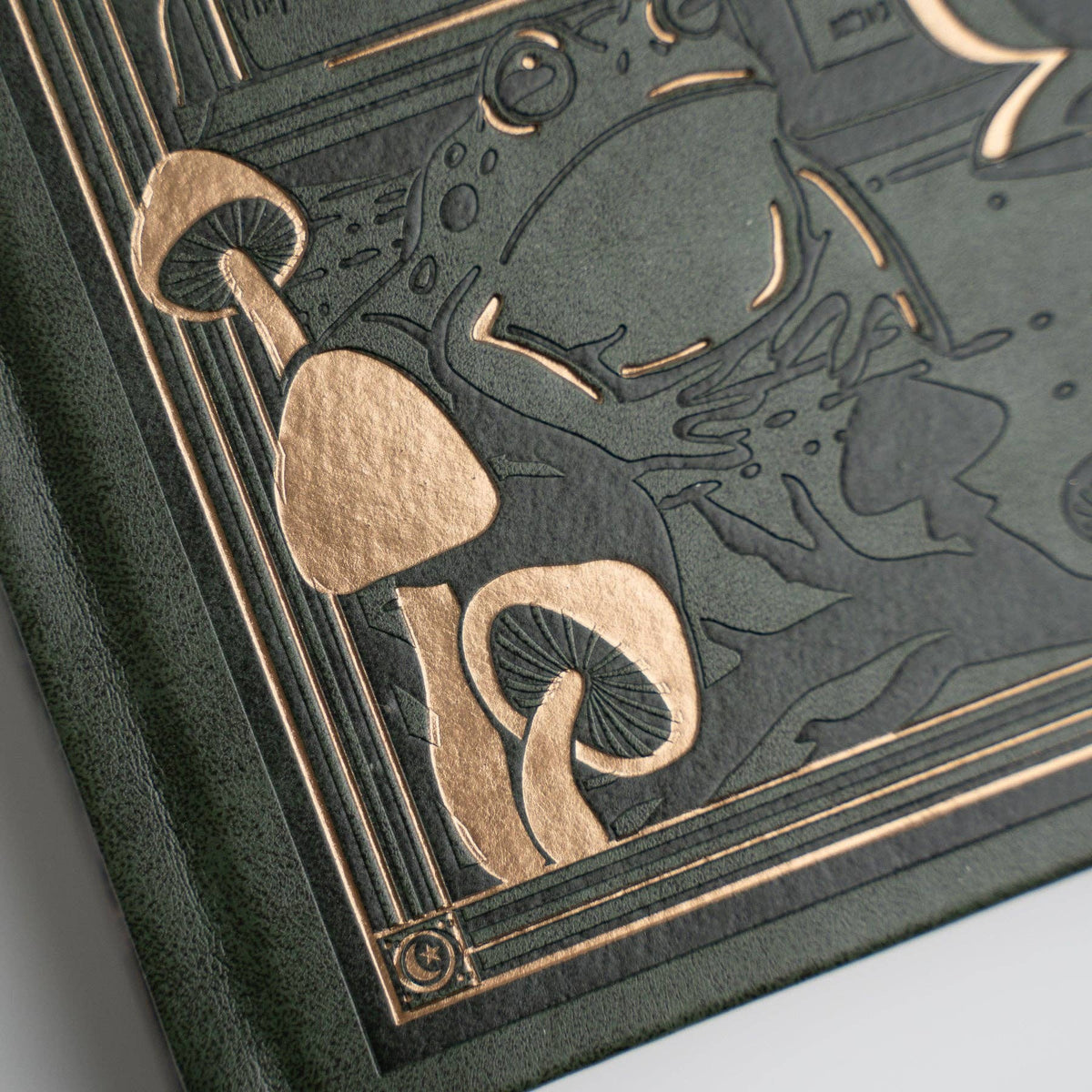 Close-up of a dark green embossed book or notebook cover with metallic gold foil detailing depicting mushrooms and abstract line art.