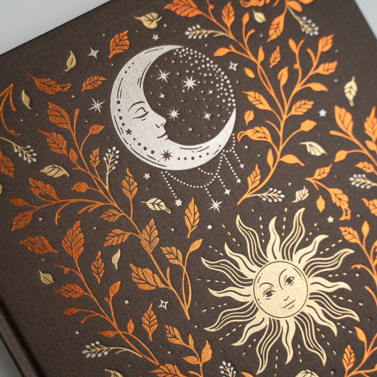 A close-up of a dark brown notebook or journal cover featuring a celestial pattern with a crescent moon, stars, a sun face, and orange and cream botanical leaves.
