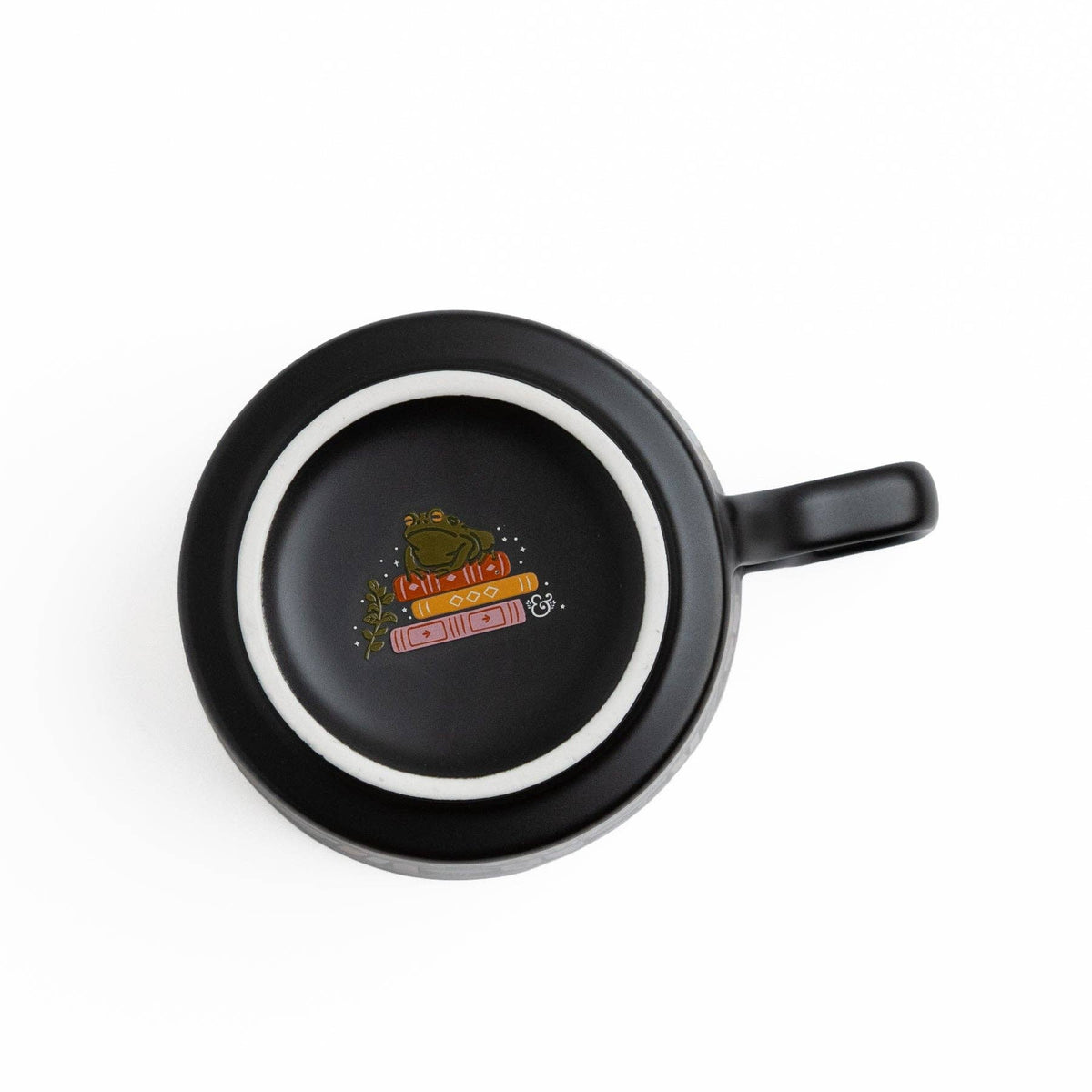 A black ceramic mug with a handle, shown from a top-down perspective on a white background. The inside of the mug features a circular white ring and a small colorful graphic in the center depicting a frog wearing a crown and holding a scroll that reads 'KTH - 11-21' and 'SWEETEST'.