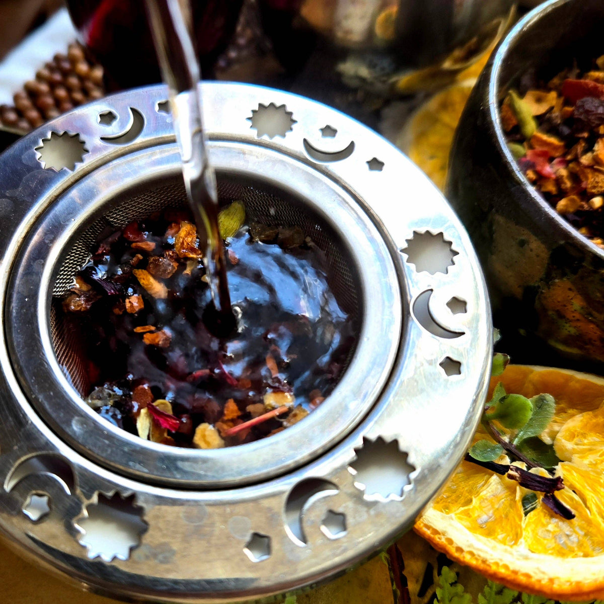 A close-up of a metal tea infuser or tea burner with a strainer basket filled with loose tea leaves and spices, with hot dark tea being poured from a metal spout. An orange slice and green leaves sit nearby on a tabletop.