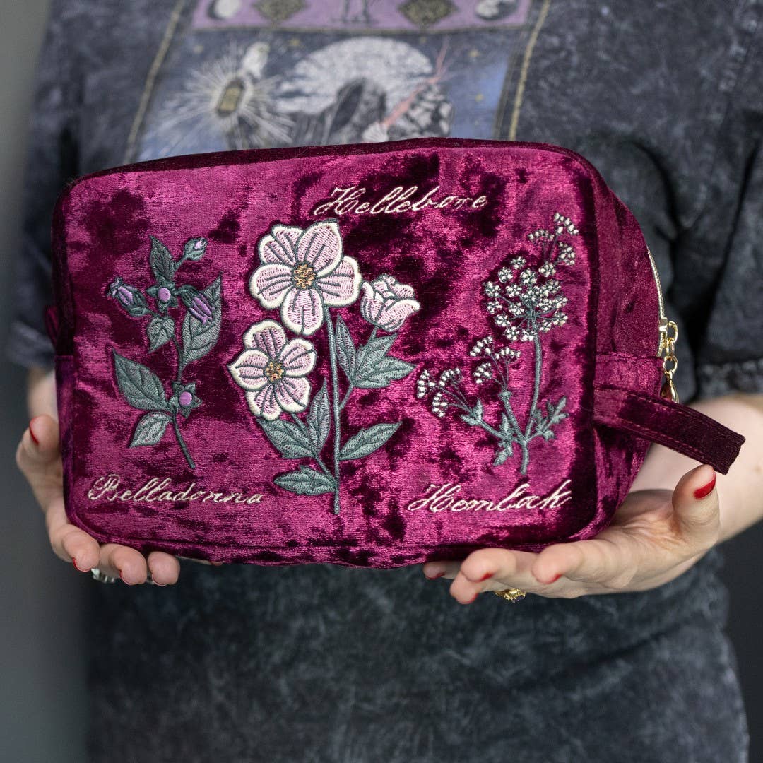 A person holds a deep magenta velvet cosmetic pouch decorated with embroidered flowers and German text labels. The person is wearing a dark gray textured shirt.