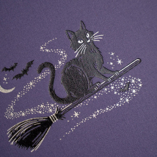 A close-up of a purple fabric featuring a black cat riding a broomstick with white star and moon embroidery details.