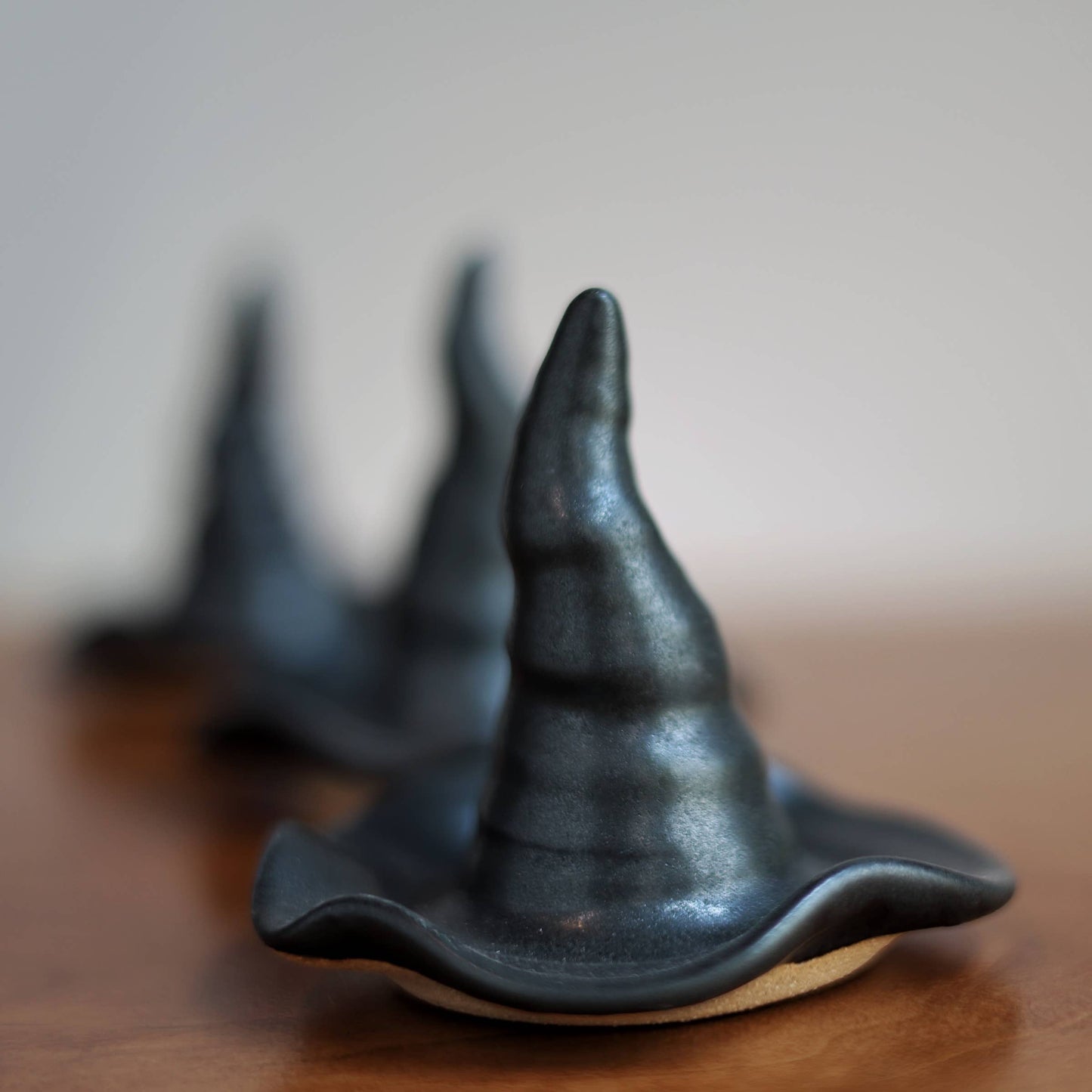 Three small black witch hat-shaped objects arranged in a row on a wooden surface, with the front one in sharp focus and the others blurred in the background against a soft neutral backdrop.