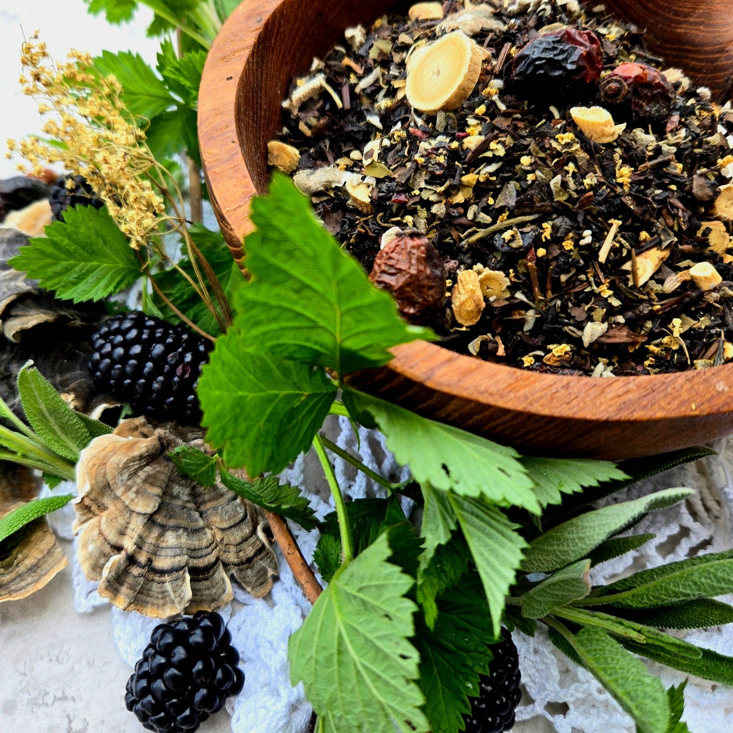 Wooden bowl filled with a mix of herbs and spices surrounded by blackberries and green leaves on a textured surface.