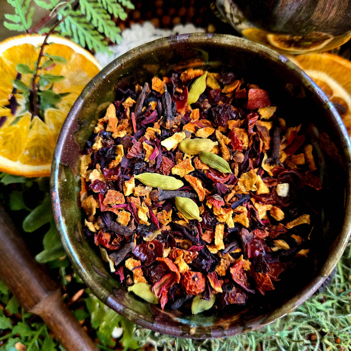 A close-up overhead shot of a dark ceramic bowl filled with loose-leaf herbal tea. The tea blend contains dried flowers, citrus peel, green cardamom pods, and whole spices.