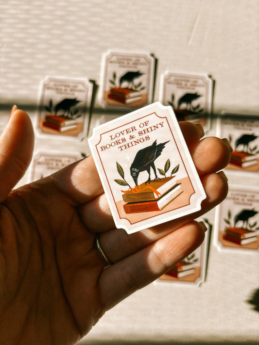 Hand holding a card with a design of a dog and text 'Lover of books & shiny things' against a blurred background.