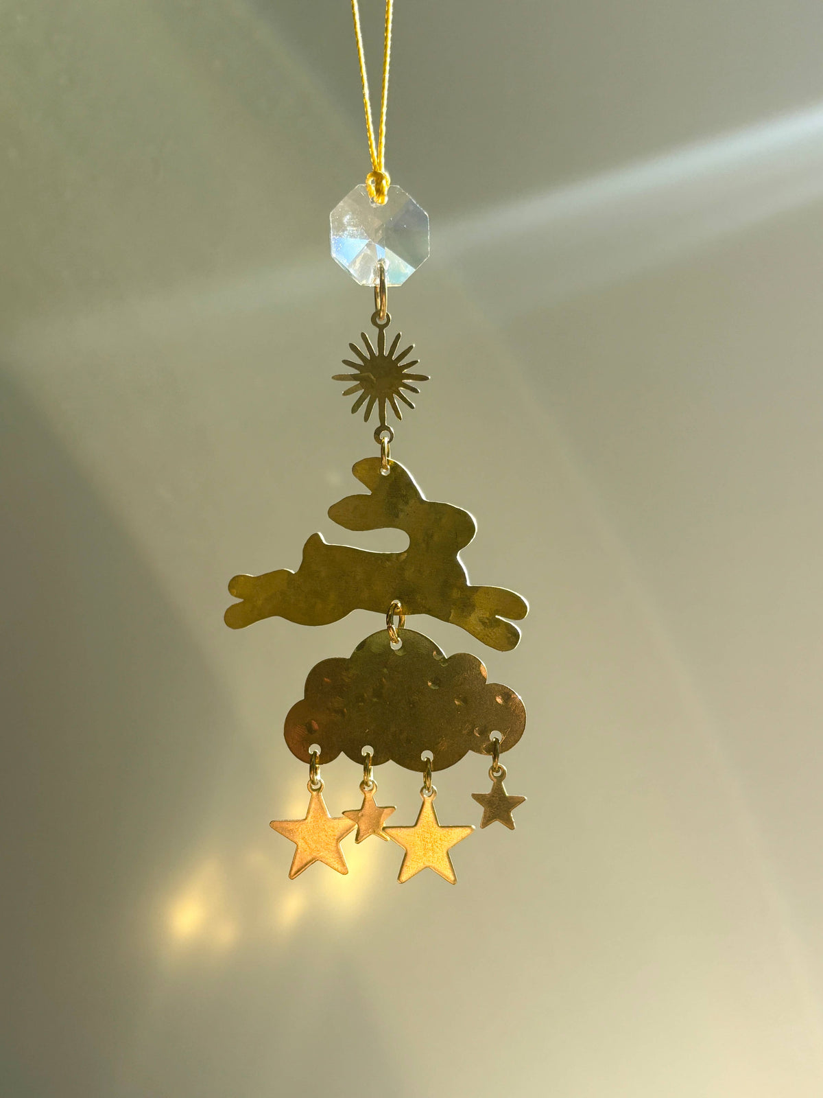 A hanging metal ornament with a faceted clear bead, gold star, sunburst, rabbit silhouette, cloud, and dangling gold stars, photographed against a softly lit neutral background.