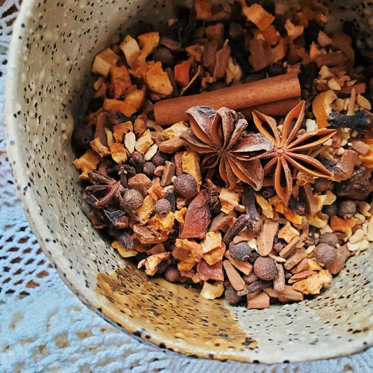 A close-up top-down view of a rustic speckled ceramic bowl filled with a warm spice blend containing cinnamon sticks, star anise, cloves, and dried citrus pieces, set on a white lace doily.