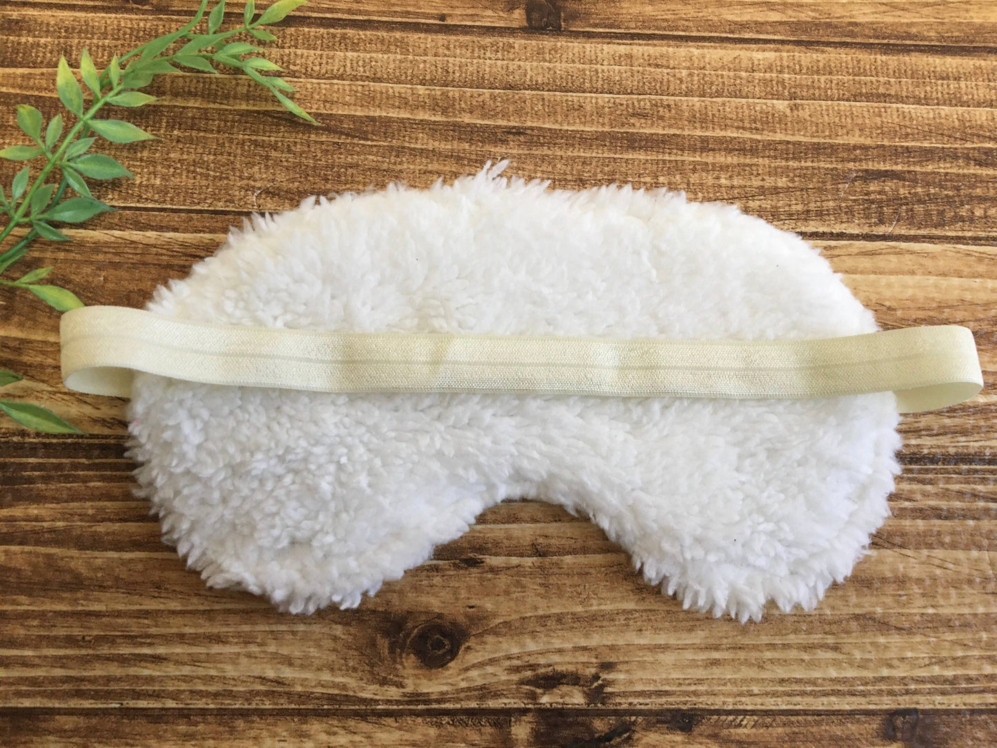 White fluffy eye mask on a wooden surface with green leaves
