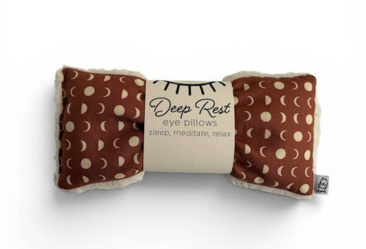 A brown eye pillow with a cream fabric trim and a repeating cream moon phase pattern, wrapped in a paper band that reads 'Deep Rest eye pillows sleep, meditate, relax'.