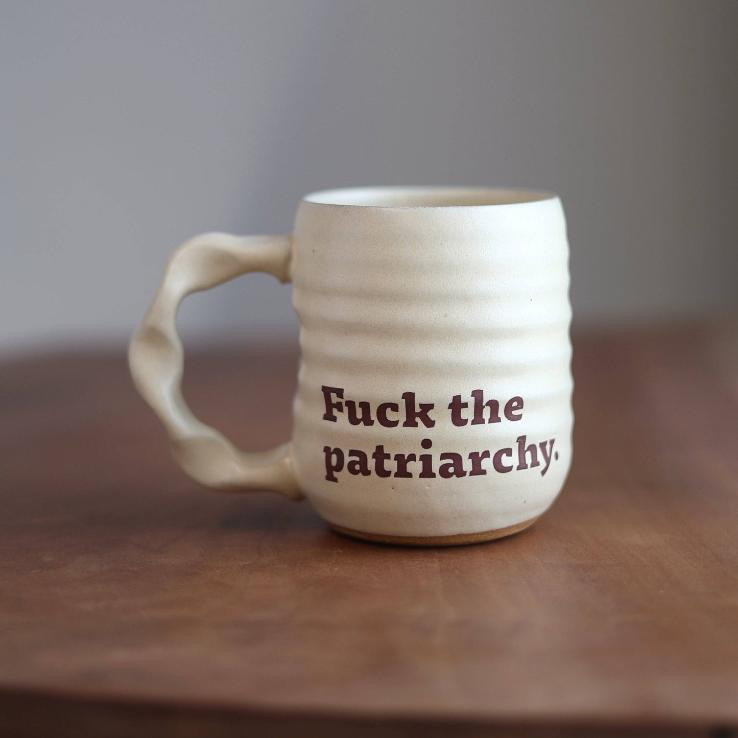 An off-white ceramic mug with a wavy handle and horizontal ridges, featuring the printed text 'Fuck the patriarchy.' in dark brown.