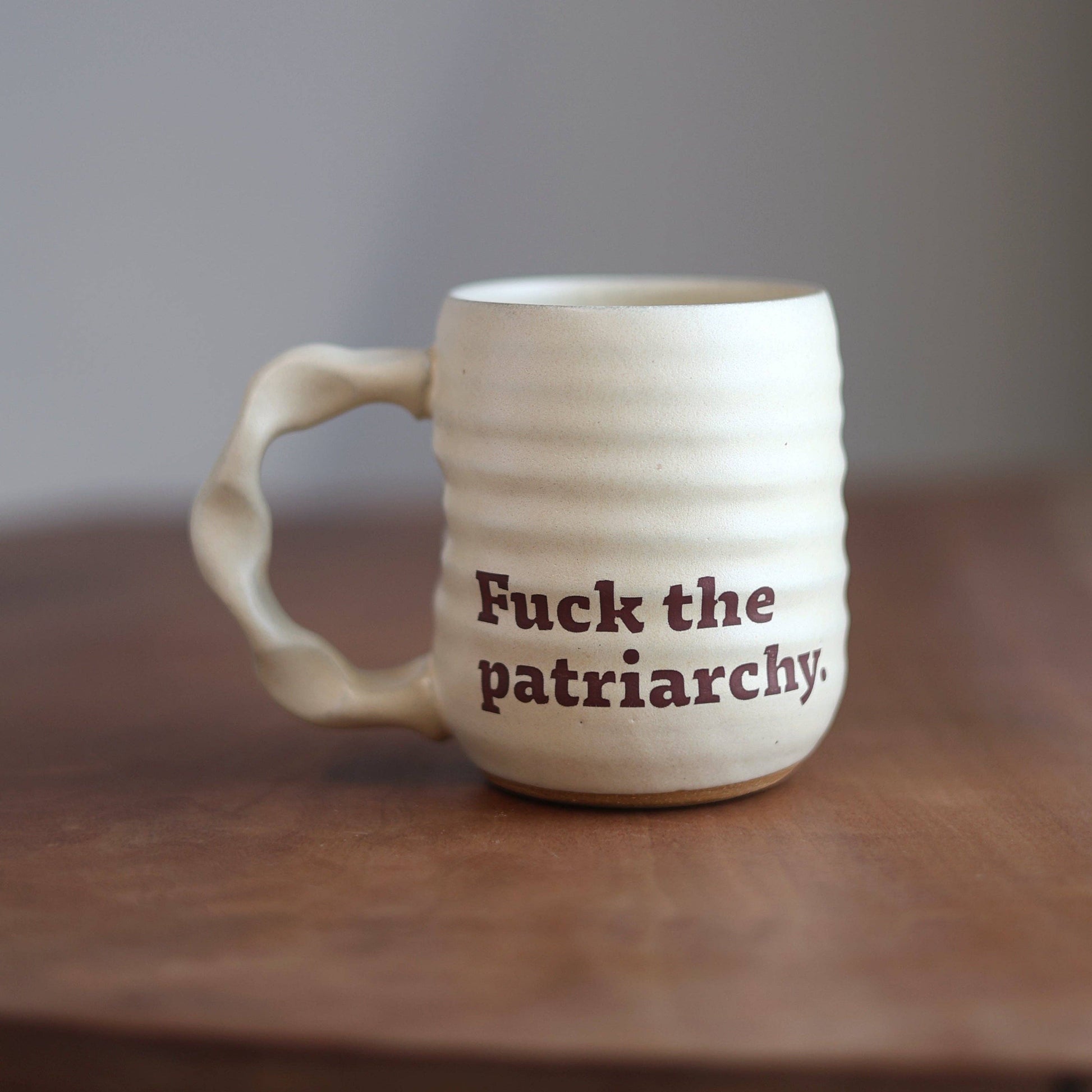 An off-white ceramic mug with a wavy handle and horizontal ridges, featuring the printed text 'Fuck the patriarchy.' in dark brown.
