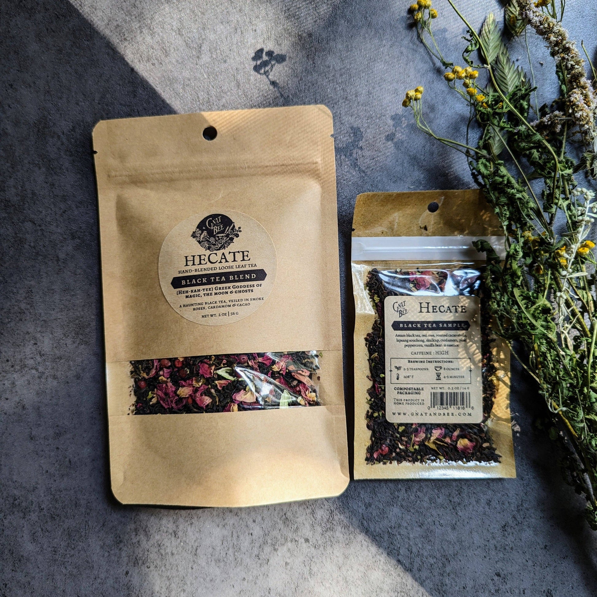 Two Hecate Black Tea Blend packages on a textured surface with dried herbs.