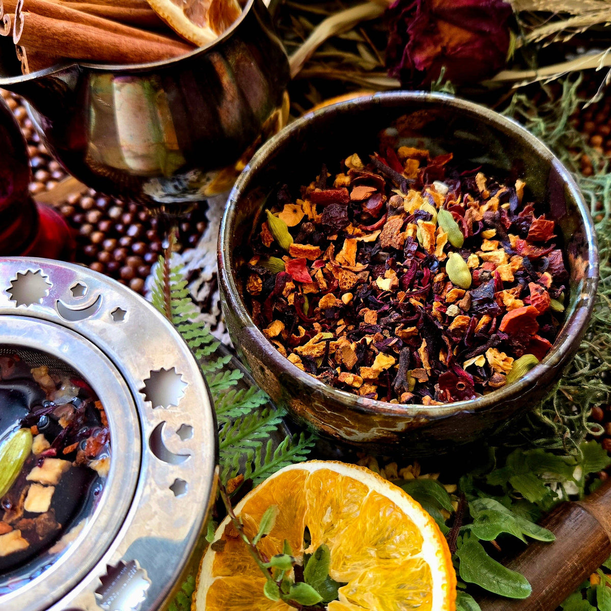 A close-up styled product shot of loose-leaf herbal tea in a rustic bowl, surrounded by cinnamon sticks, green leaves, a dried rose, and a sliced orange. A metal tea infuser and part of a metal vessel are visible at the edges.
