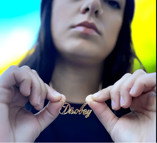 Gold necklace with 'Disobey' pendant held by hands against a blurred background