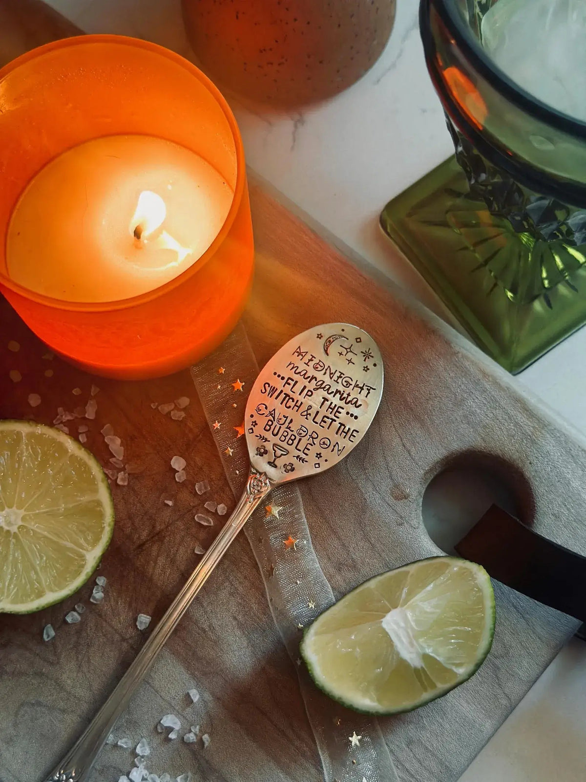 A silver spoon engraved with a whimsical quote about moonlight and bubbles, styled on a wooden board with a lit orange candle, lime wedges, and a green glass bowl.