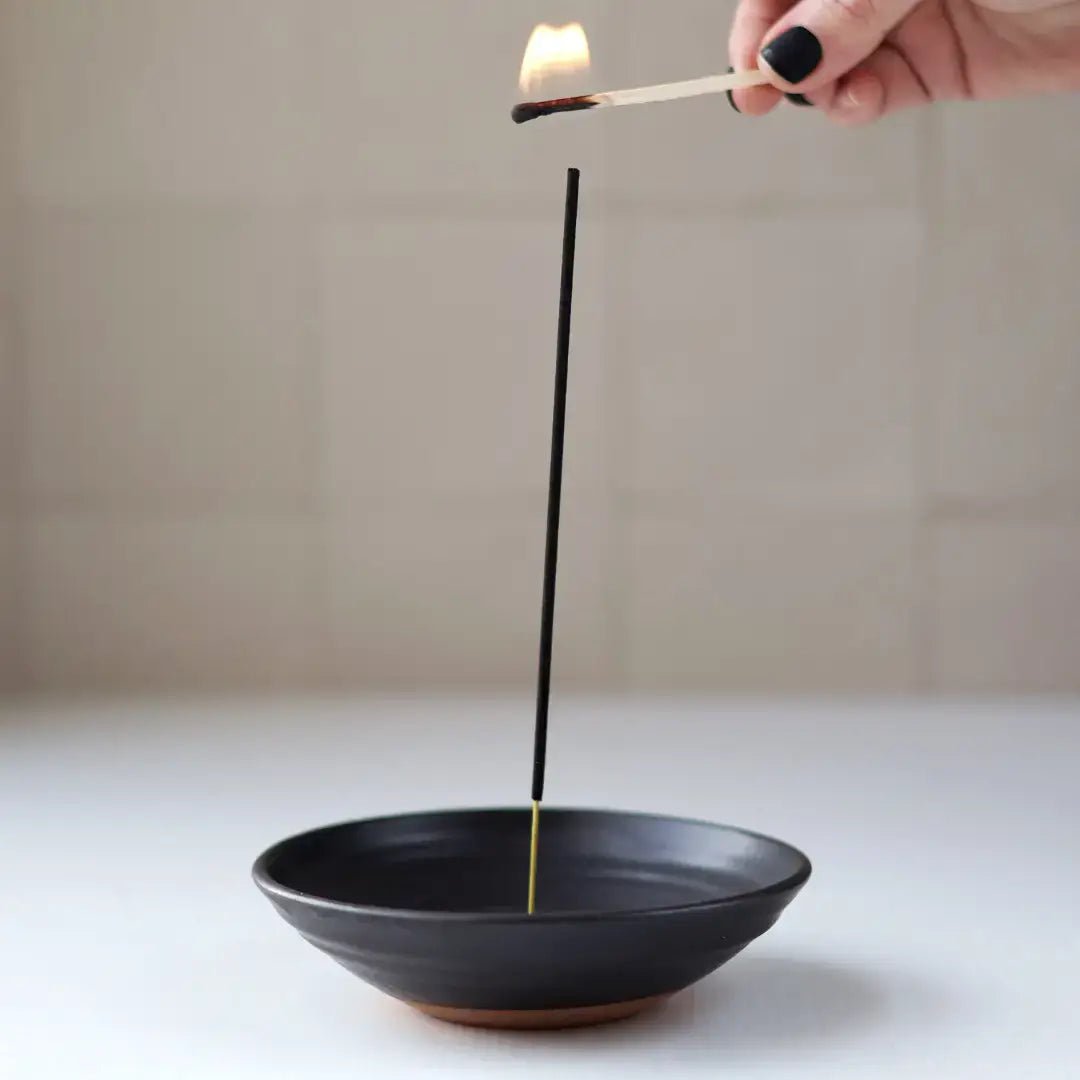 Incense stick being lit over a black incense bowl on a neutral background