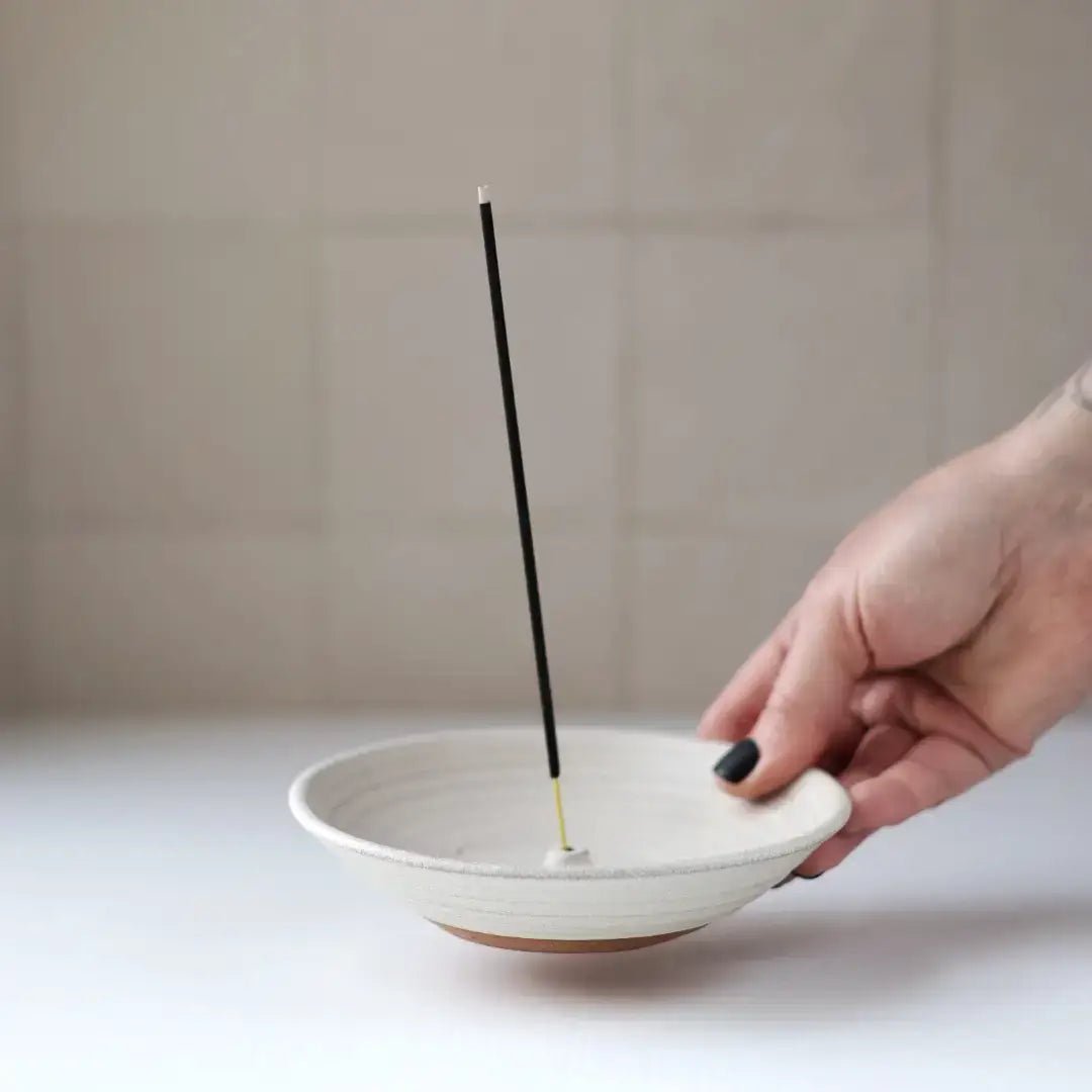 Hand placing an incense stick into a white ceramic bowl on a neutral background