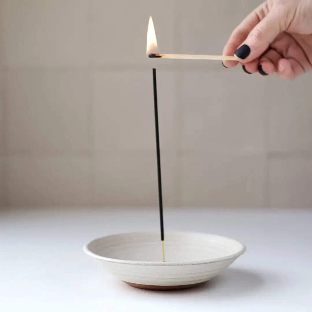 Person lighting an incense stick with a match over a white dish on a neutral background